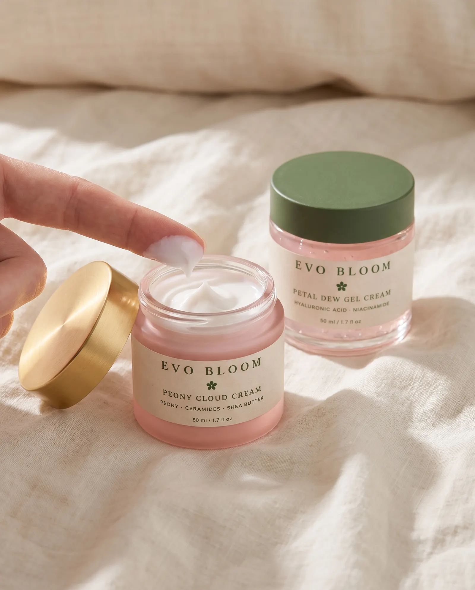 A fingertip lifting cream from an open jar of Peony Cloud Cream, with Petal Dew Gel Cream on the linen behind